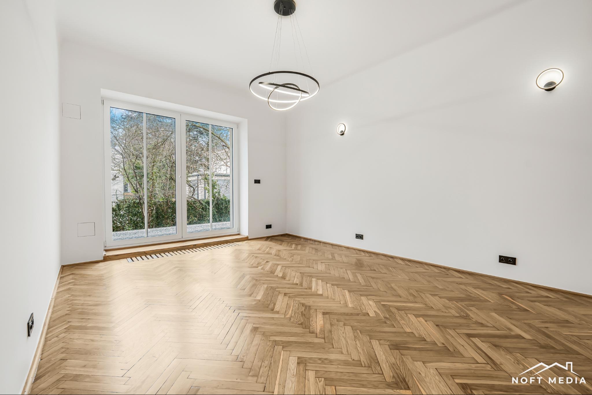 Pusty pokój z parkietem ułożonym w jodełkę, nowoczesną lampą sufitową i oknem balkonowym z widokiem na zieleń.