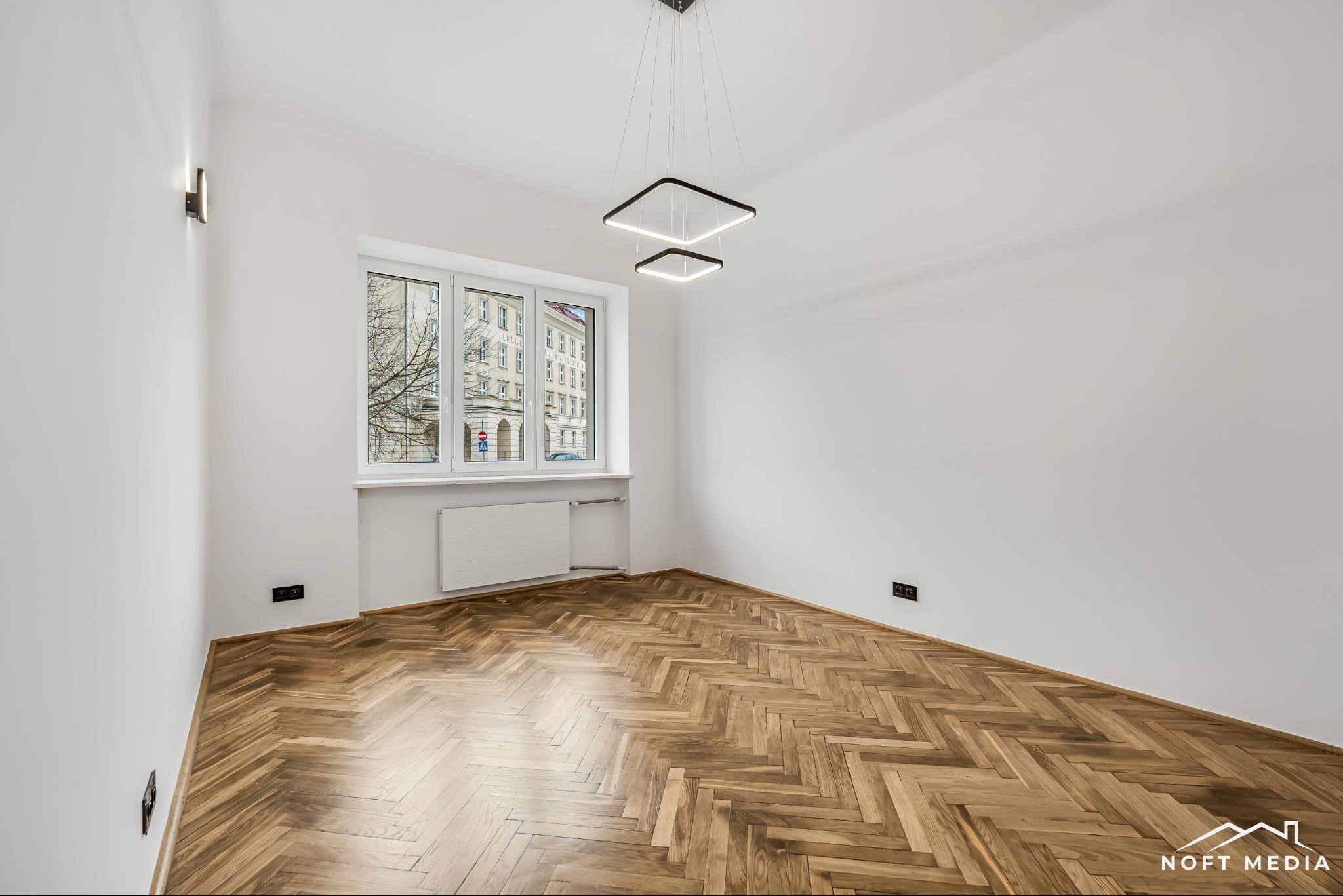 Puste, jasne wnętrze z parkietem w jodełkę, nowoczesnym oświetleniem w postaci dwóch kwadratowych lamp wiszących, białymi ścianami i oknem z widokiem na budynek z czerwonej cegły.