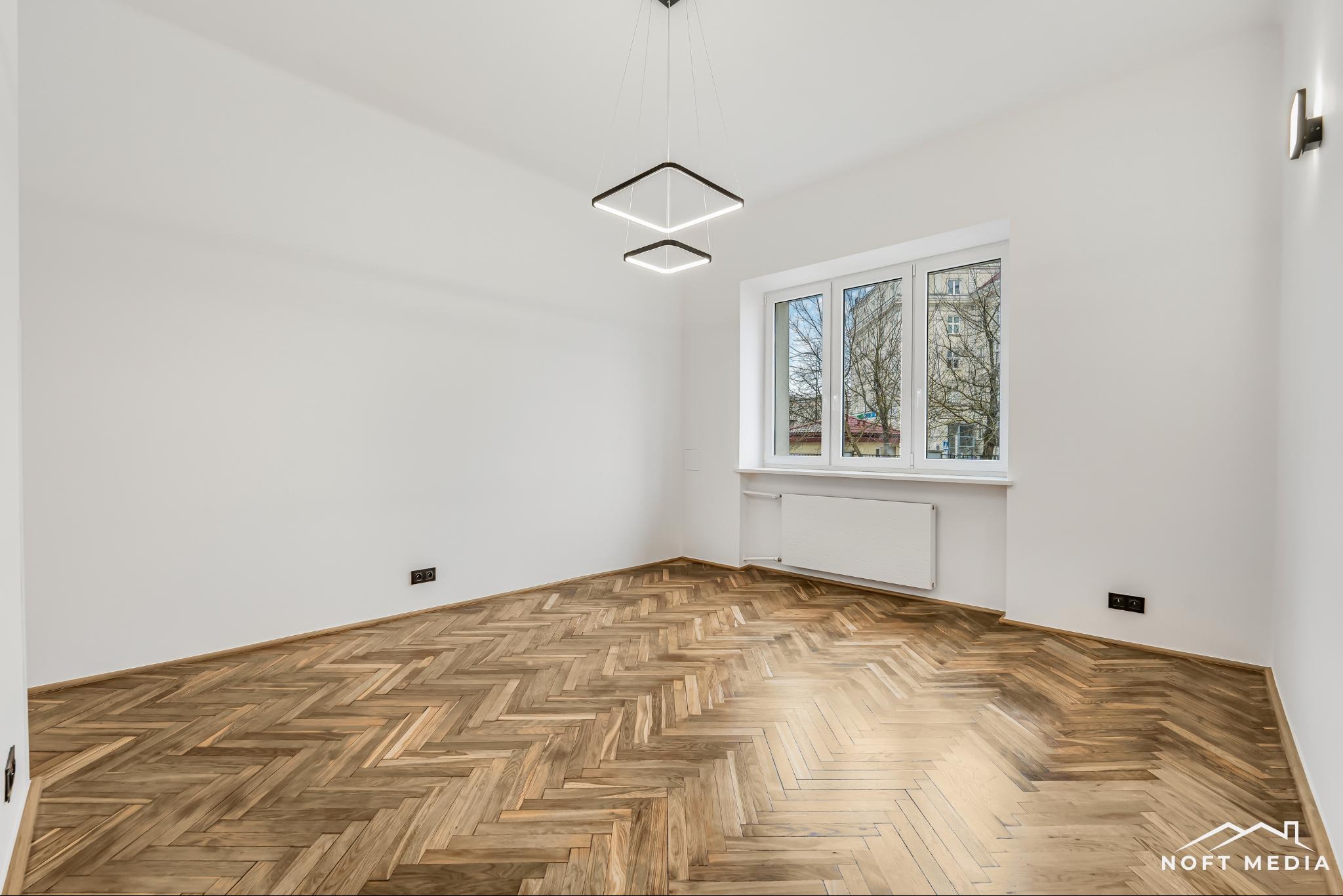 Puste, jasne wnętrze z parkietem w jodełkę, nowoczesnym oświetleniem w kształcie kwadratów i widokiem na budynek przez okno.