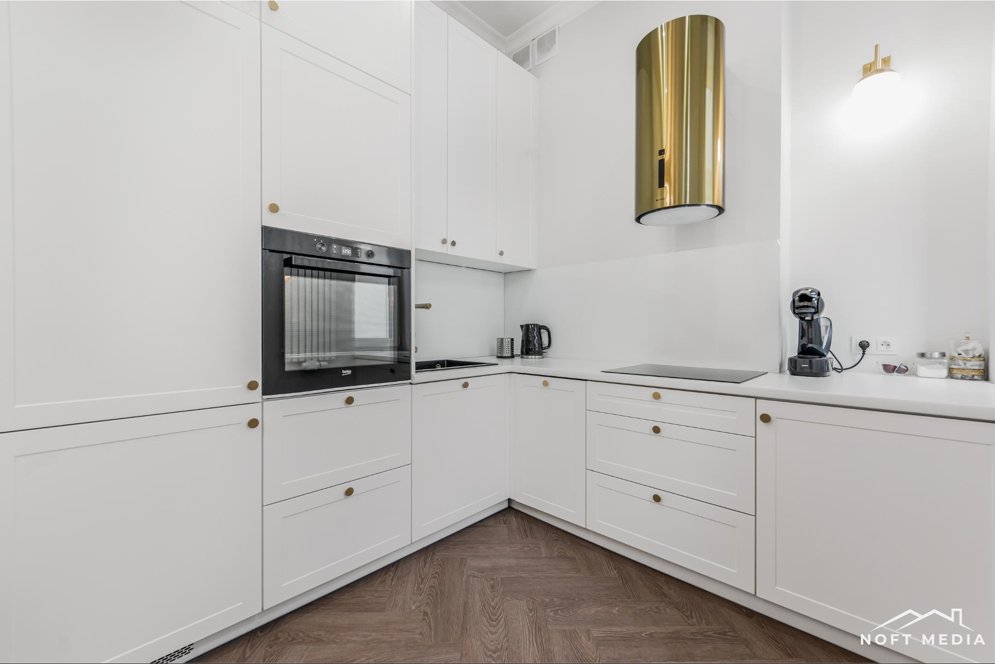 Biała kuchnia z parkietem ułożonym w jodełkę, złoty okap cylindryczny, sprzęt AGD w zabudowie, minimalistyczne uchwyty.