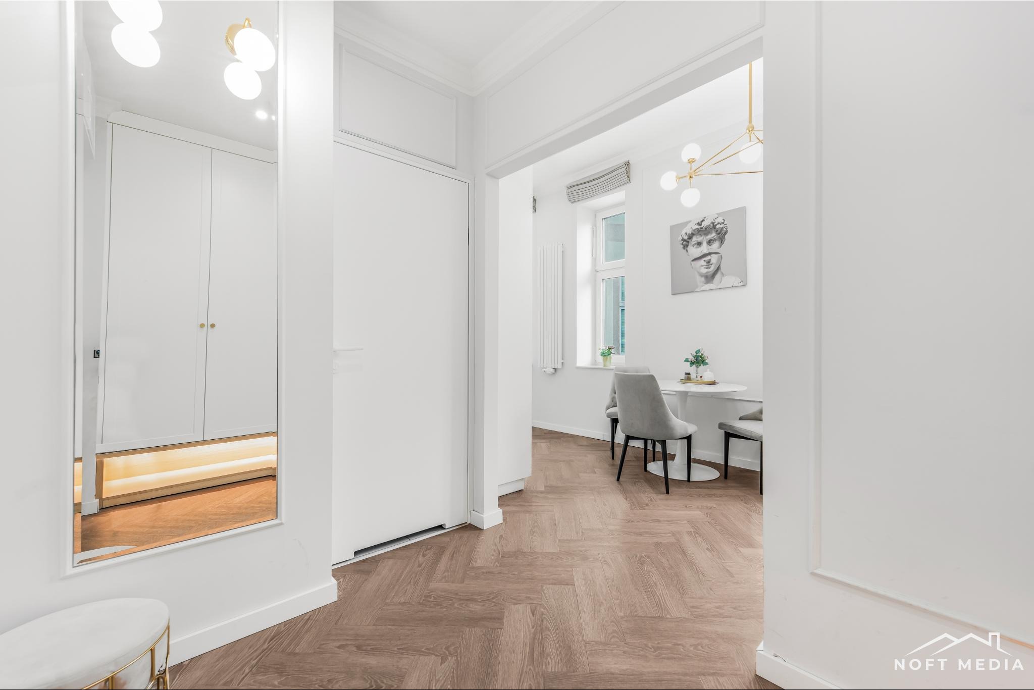 Jasne wnętrze mieszkania z parkietem w jodełkę, widok na jadalnię z okrągłym stołem i szarymi krzesłami, lustro odbijające szafę w przedpokoju, minimalistyczny design.