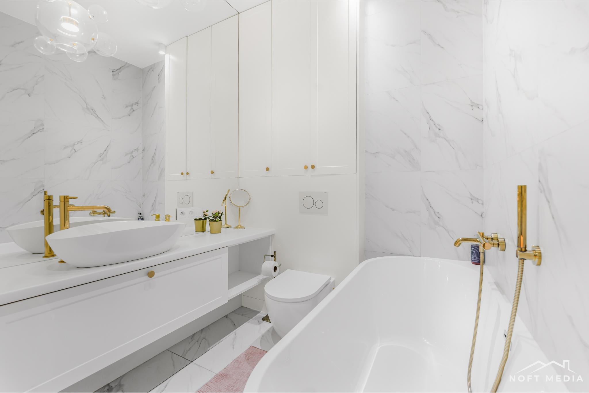 Nowoczesna, biała łazienka z marmurowymi płytkami, złotą armaturą i designerską lampą sufitową. Umywalka nablatowa o owalnym kształcie, minimalistyczna toaleta i wanna wolnostojąca tworzą spójną...