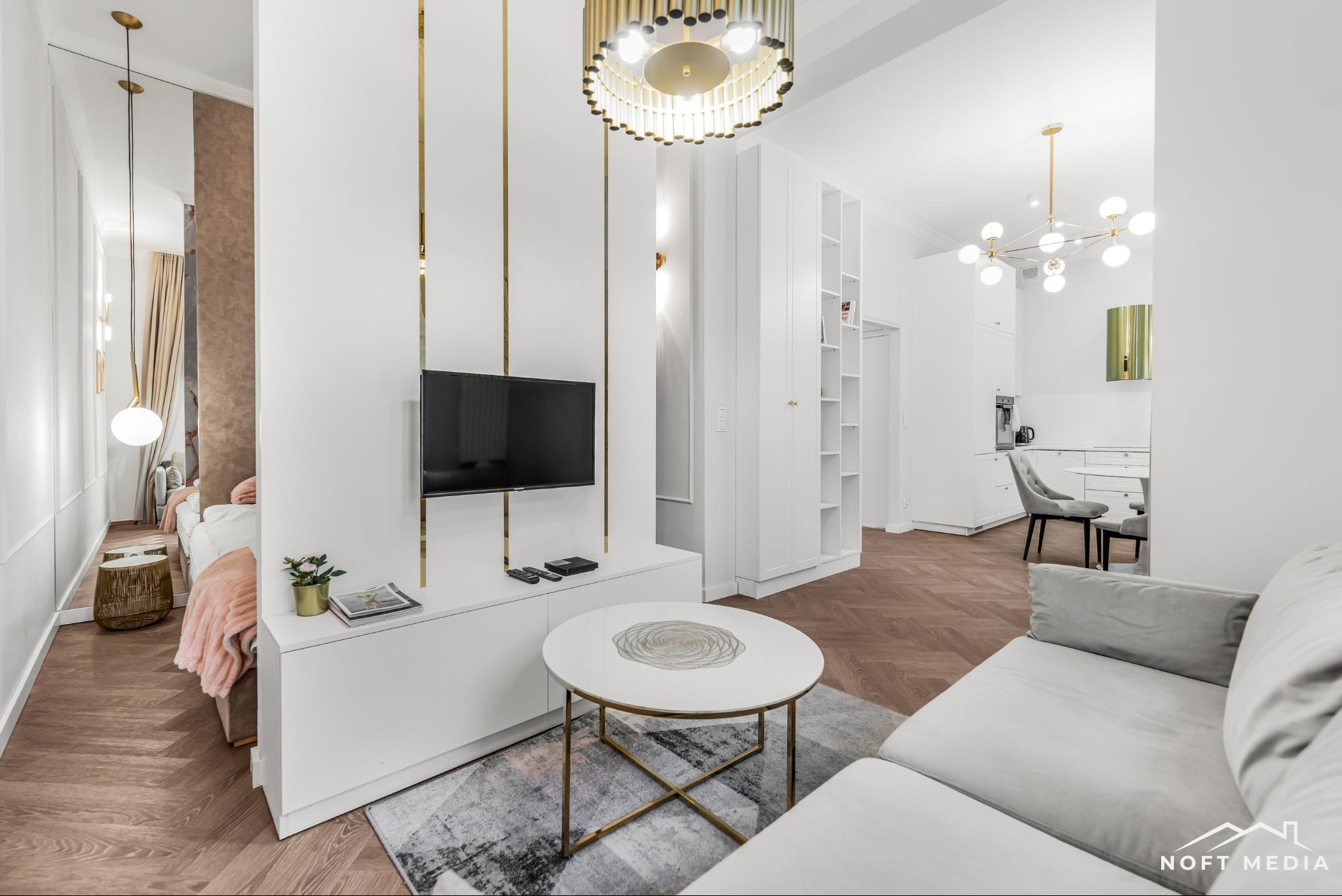 Nowoczesne wnętrze apartamentu z parkietem w jodełkę, białymi meblami, złotymi akcentami i dekoracyjnym oświetleniem, widok na salon, sypialnię i kuchnię.
