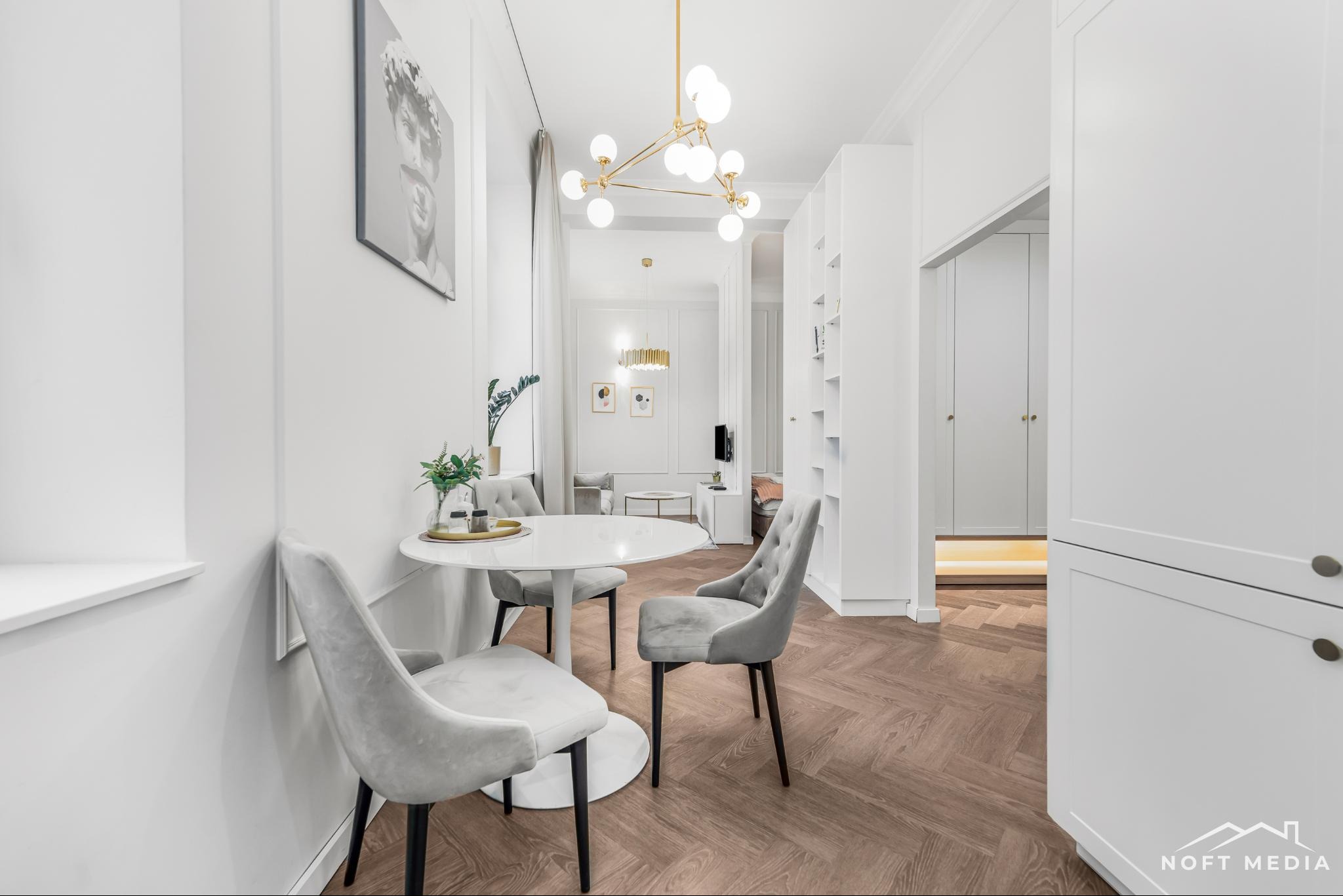 Jasne wnętrze jadalni z okrągłym stołem, szarymi krzesłami i parkietem w jodełkę, widok na salon z dekoracyjnym oświetleniem i minimalistycznymi obrazami na ścianie.