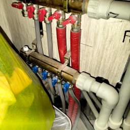Ruslan Skalichuk - Rozdzielacz ogrzewania podłogowego z zaworami termostatycznymi, rurami grzewczymi w izolacji czerwonej, oraz elementami instalacji wodnej z tworzywa sztucznego.