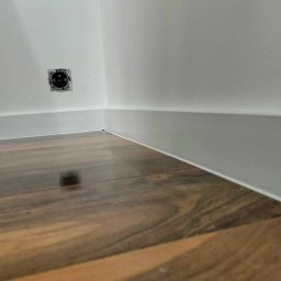 Ruslan Skalichuk - Ukończone połączenie ściany z podłogą: biała listwa przypodłogowa przy brązowych panelach, widoczne gniazdko elektryczne w ścianie.