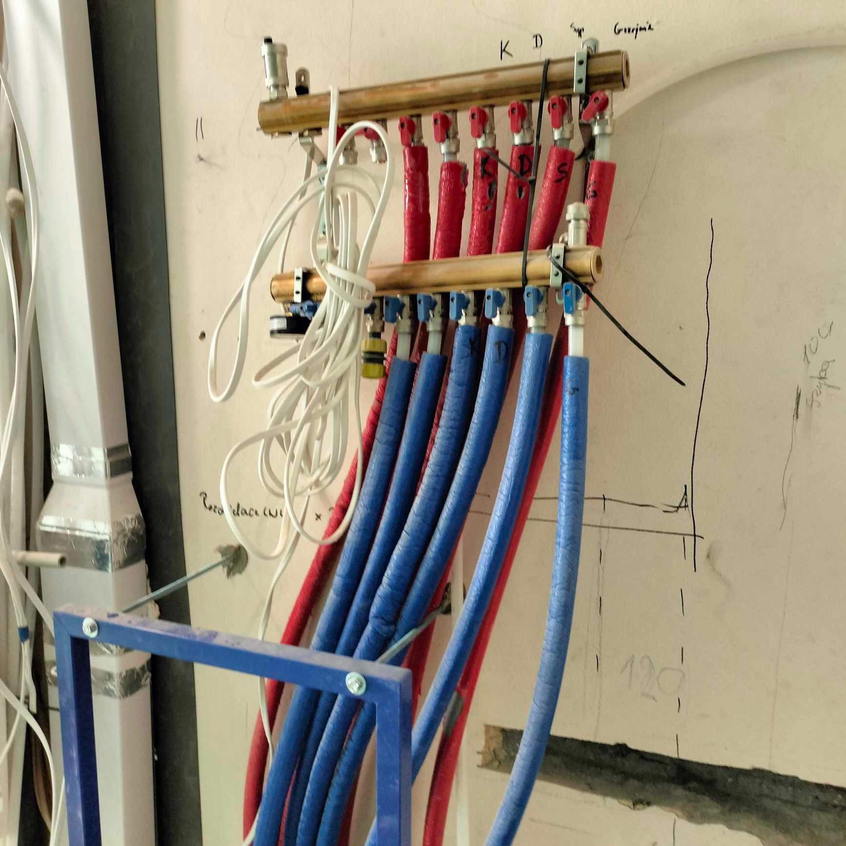 Rozdzielacz ogrzewania podłogowego z rurami w kolorze czerwonym i niebieskim, zamontowany na ścianie z widocznymi odręcznymi notatkami i szkicem instalacji, na pierwszym planie fragment konstrukcji...