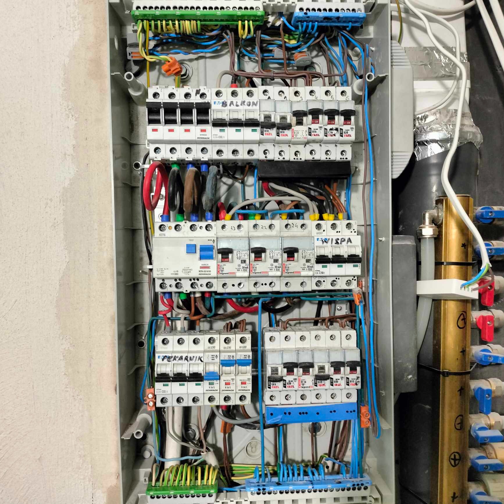 Otwarta skrzynka elektryczna z bezpiecznikami i licznymi przewodami, z widocznymi oznaczeniami 'BALKON', 'WISPA' i 'PIEKARNIK' na poszczególnych wyłącznikach, obok rury z zaworami.