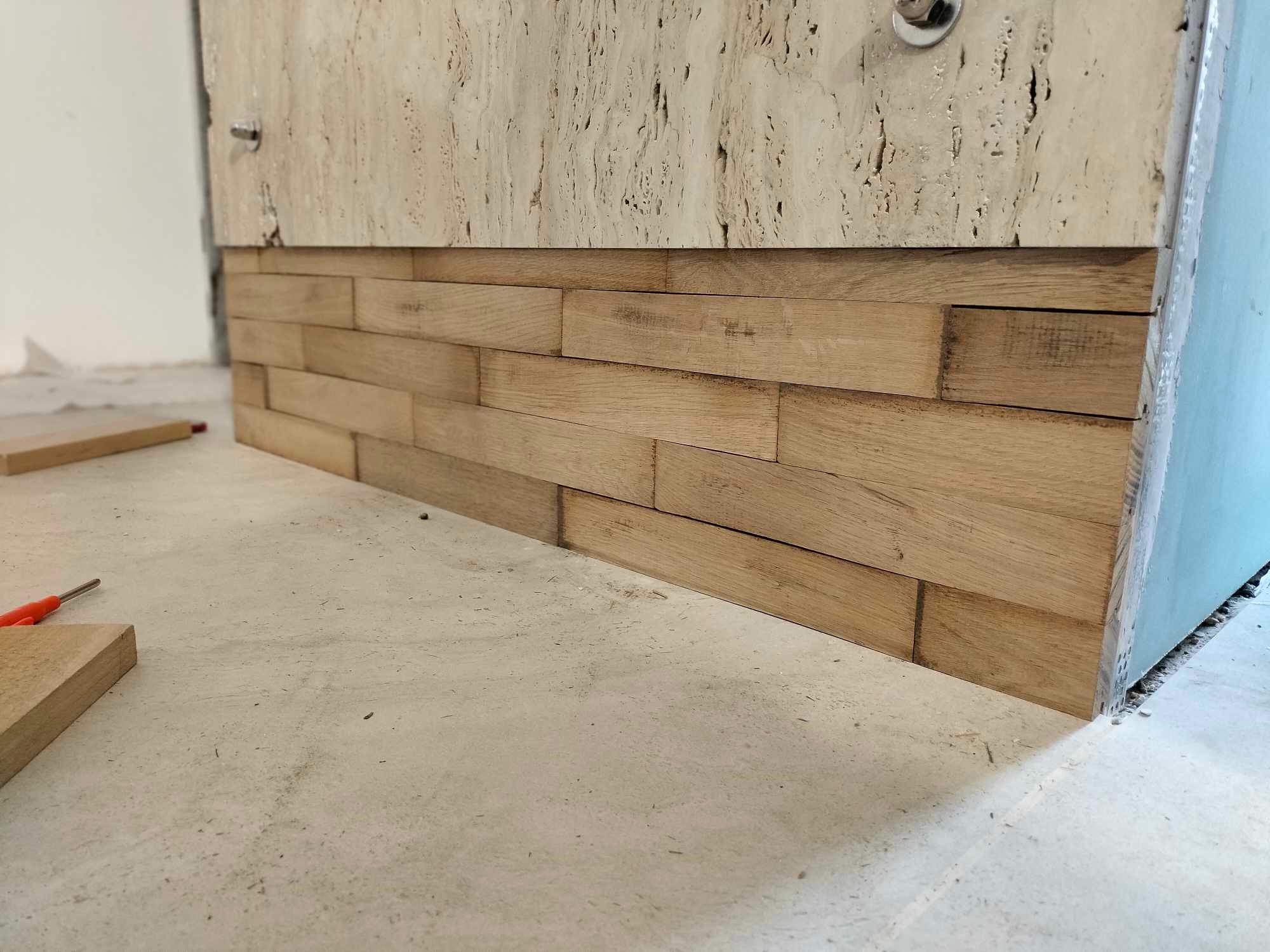 Fragment ściany z dekoracyjnymi panelami drewnianymi ułożonymi w cegiełkę, widoczne niedokończone prace i surowy beton na podłodze.