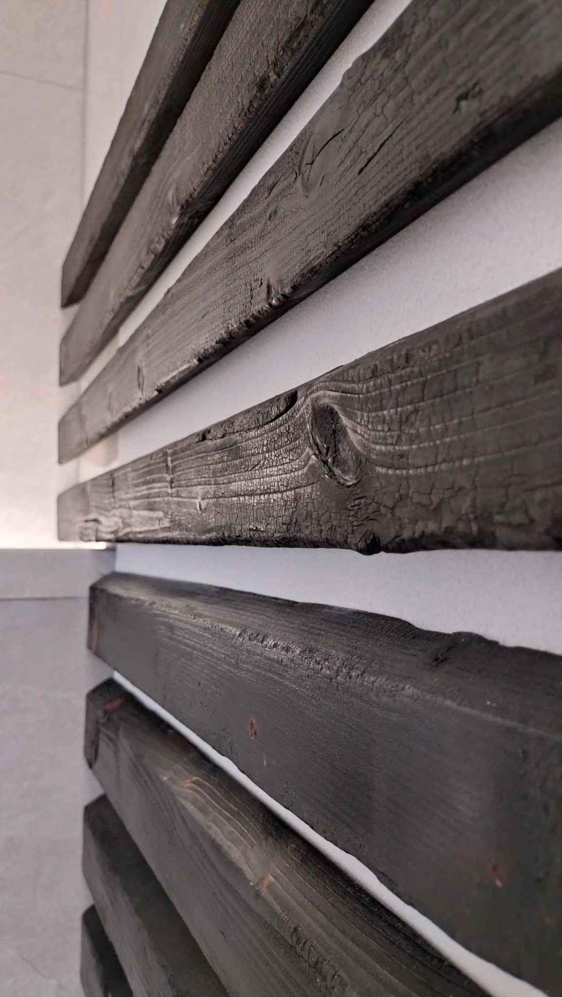 Dekoracyjne, poziome lamele z ciemnego, opalizowanego drewna na białej ścianie, z widoczną strukturą drewna i sękami, oświetlone z góry.