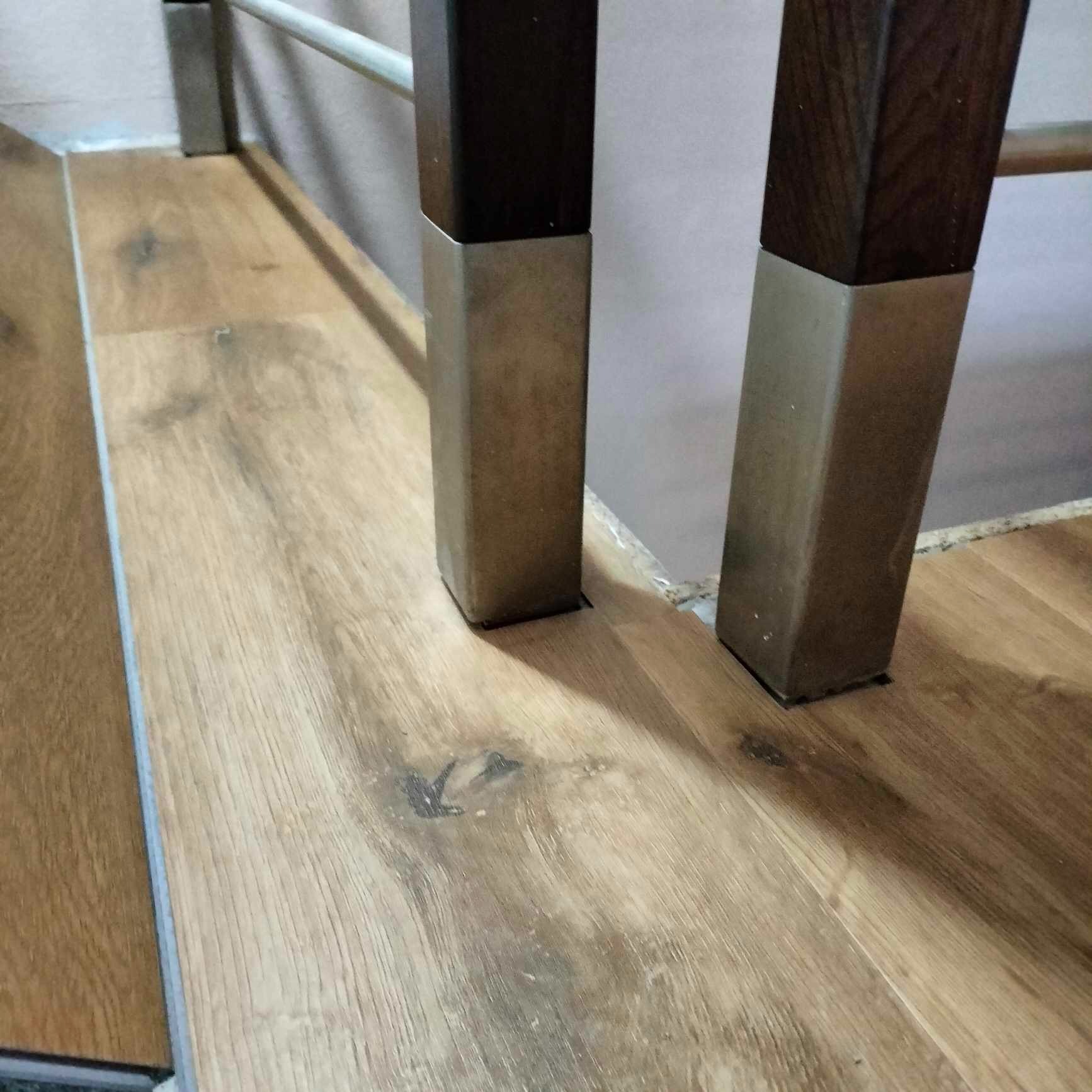 Fragment podłogi z paneli drewnopodobnych w odcieniach brązu z widocznymi nogami meblowymi z ciemnego drewna i metalu.