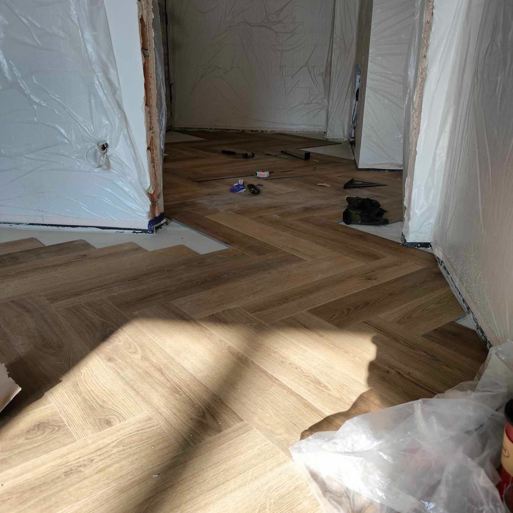 Montaż paneli podłogowych w jodełkę, zabezpieczone folią ściany, widoczne narzędzia i niedokończone fragmenty podłogi.