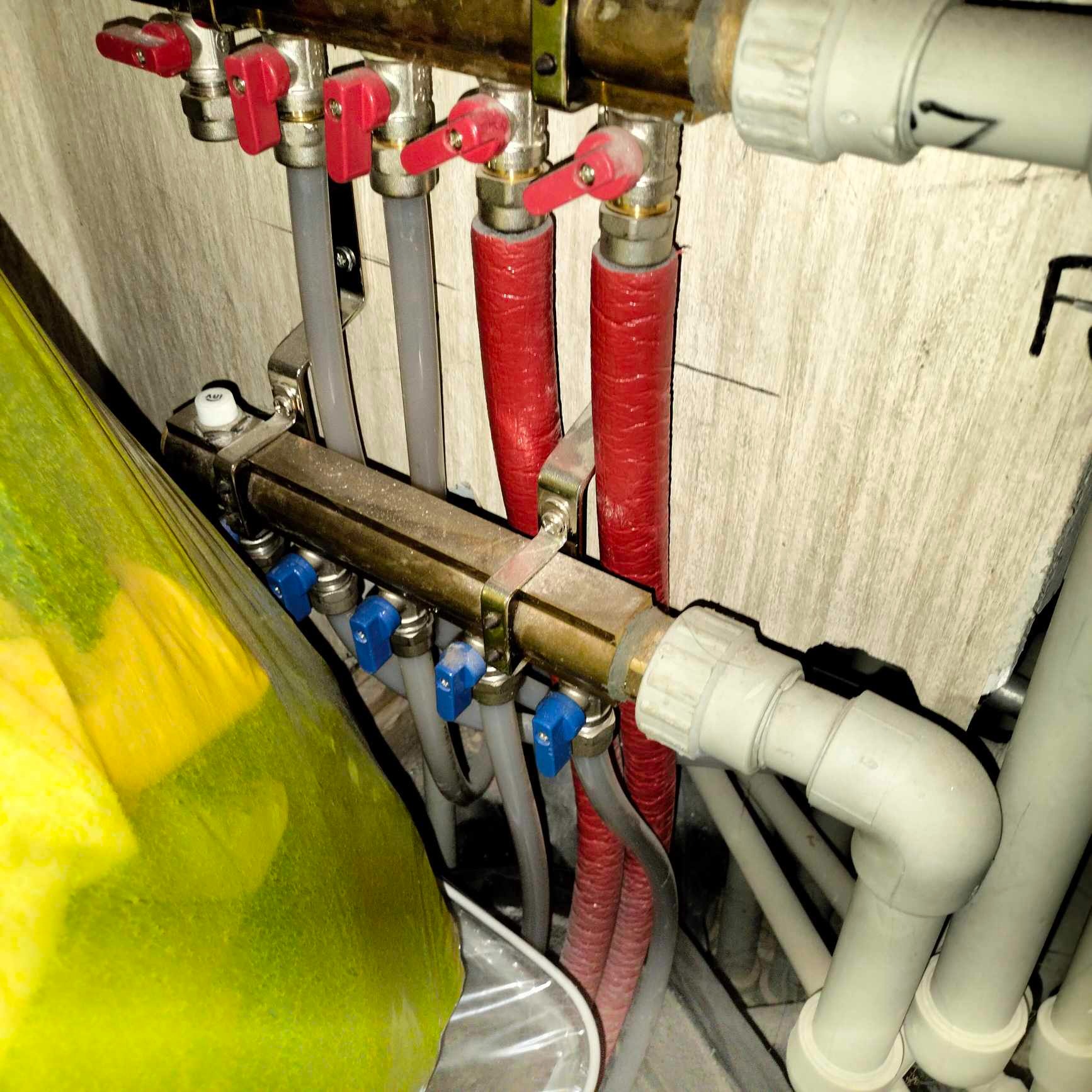 Rozdzielacz ogrzewania podłogowego z zaworami termostatycznymi, rurami grzewczymi w izolacji czerwonej, oraz elementami instalacji wodnej z tworzywa sztucznego.