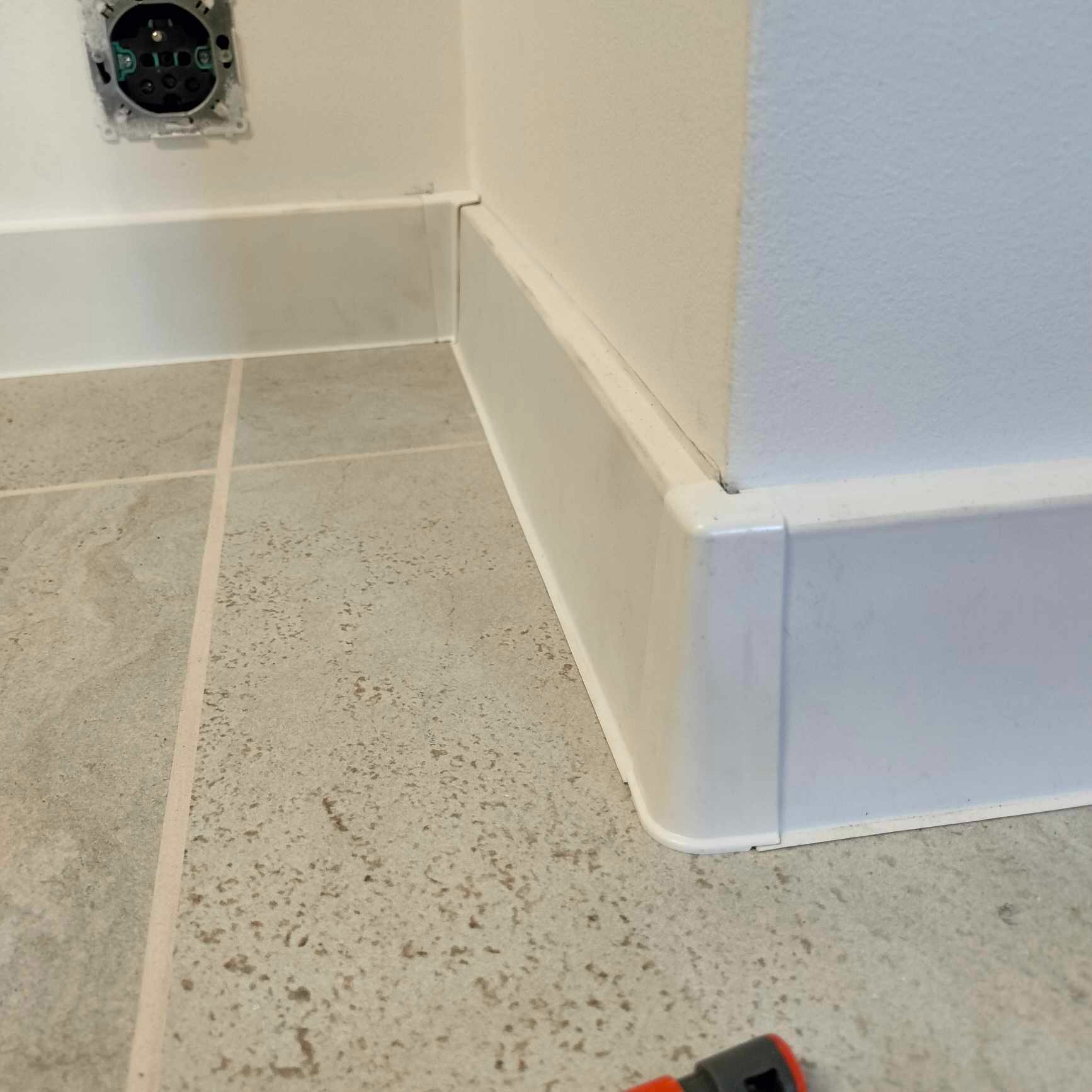 Szczegółowe ujęcie narożnika ściany z zamontowaną białą listwą przypodłogową, widoczna szara płytka podłogowa i gniazdko elektryczne bez obudowy, fragment narzędzia w rogu kadru.