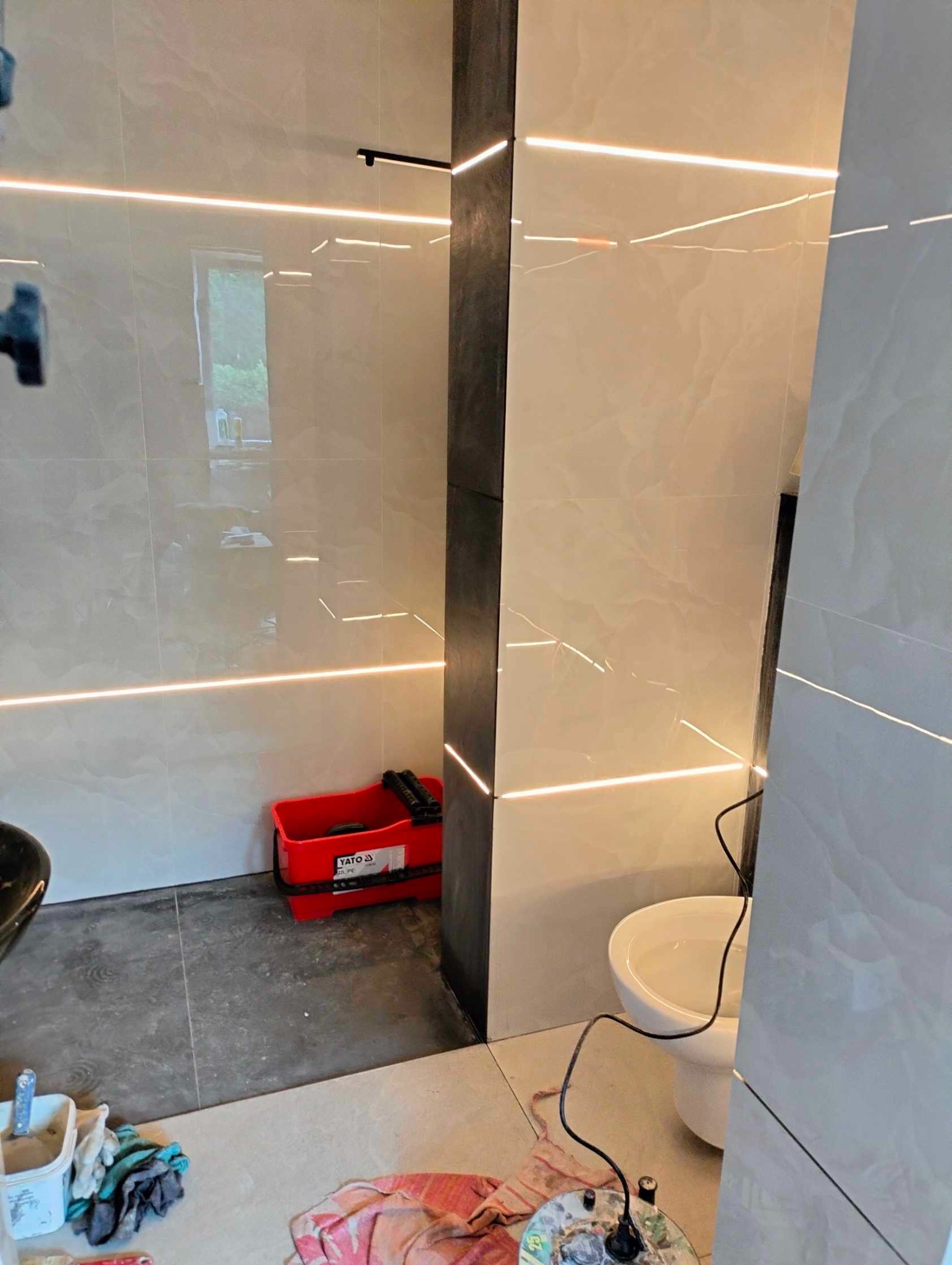 Wykończona łazienka z podświetlanymi płytkami, widoczny fragment toalety, czerwone wiadro Yato i akcesoria malarskie na podłodze.