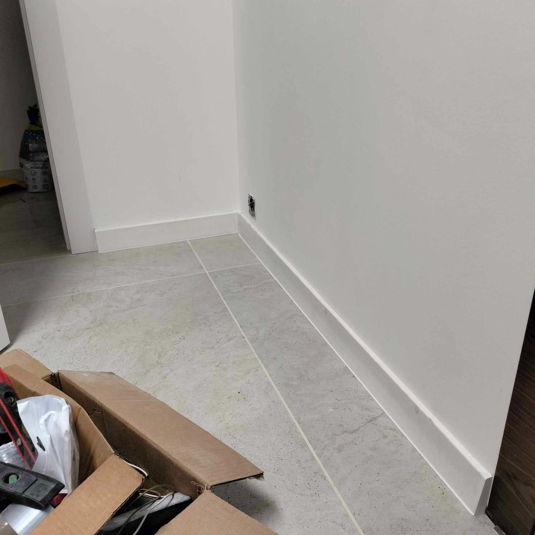 Świeżo pomalowane ściany w odcieniach bieli z zamontowanymi białymi listwami przypodłogowymi, widoczna puszka elektryczna i karton z narzędziami.