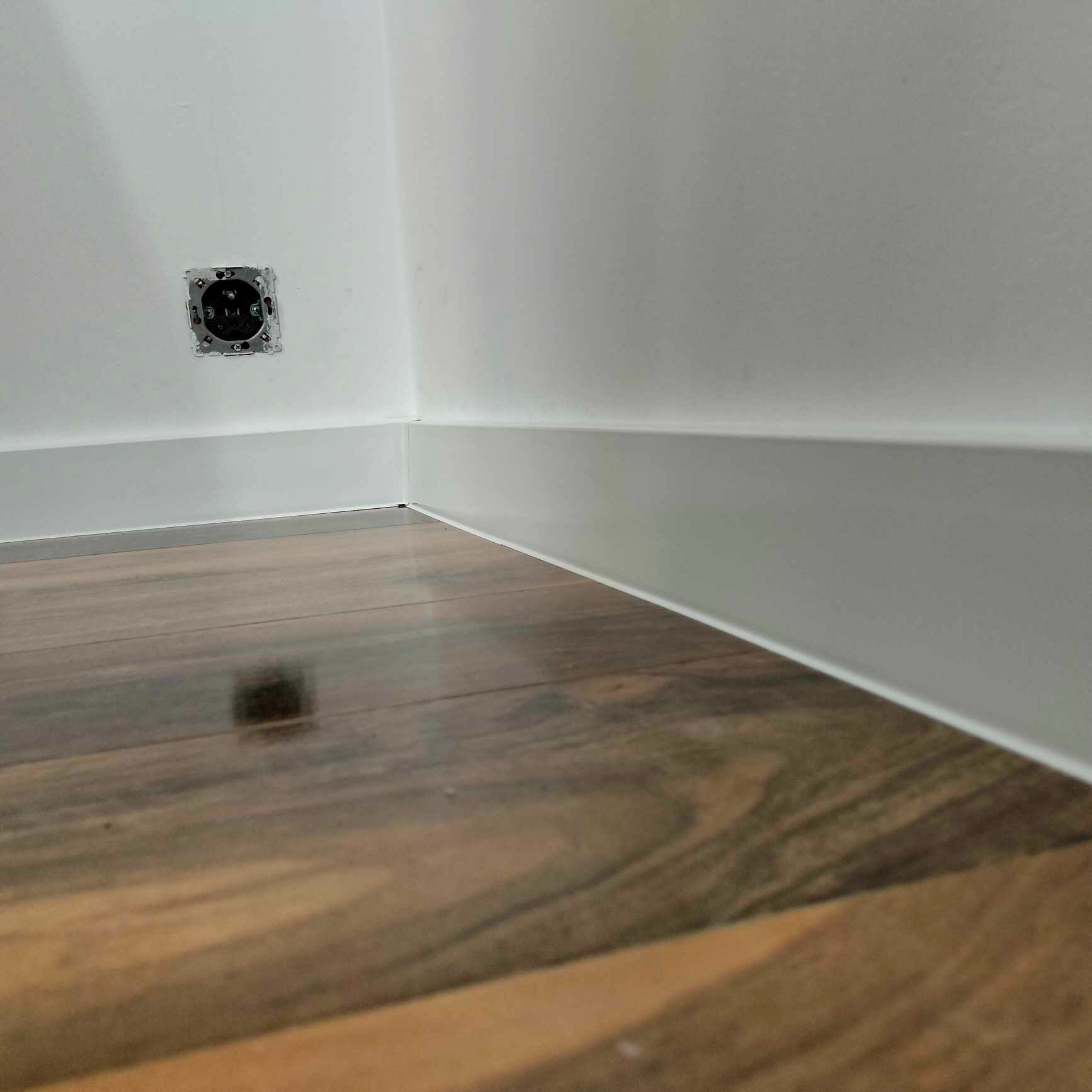 Ukończone połączenie ściany z podłogą: biała listwa przypodłogowa przy brązowych panelach, widoczne gniazdko elektryczne w ścianie.