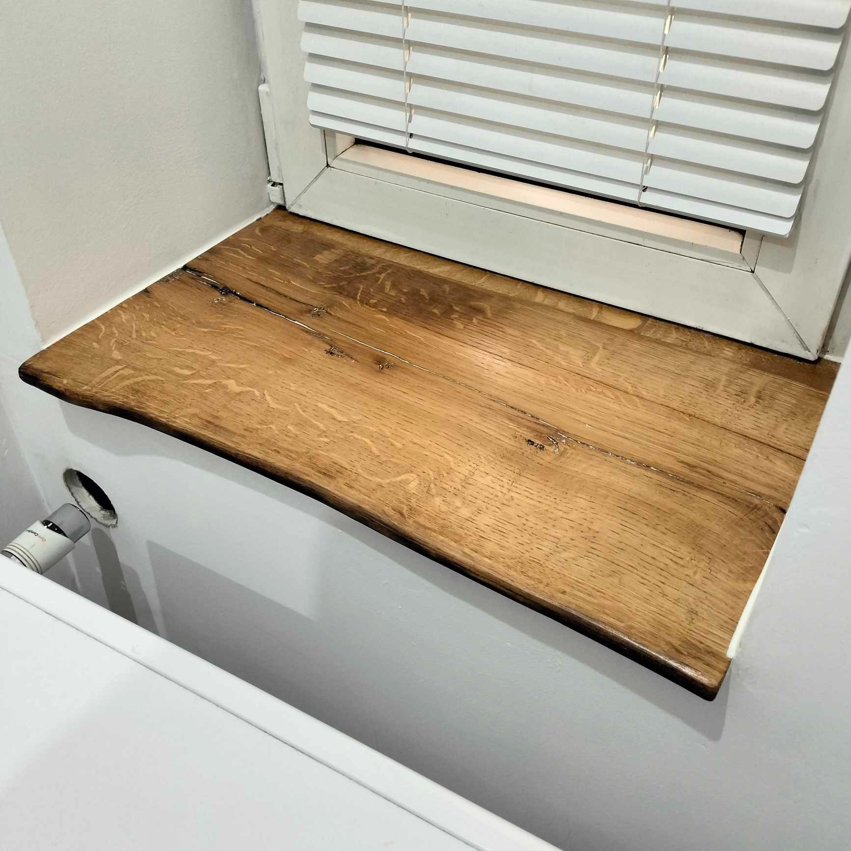 Blat z drewna dębowego, zamontowany pod oknem z żaluzjami w łazience. Widoczny fragment wanny i instalacja wodna. Naturalne wykończenie krawędzi blatu.