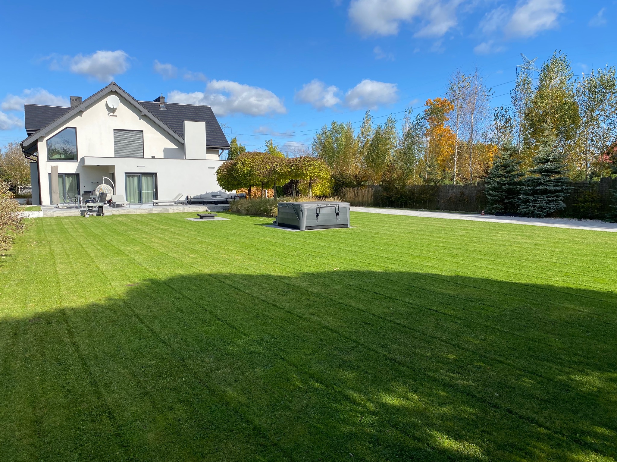 Starannie przystrzyżony trawnik z wyraźnymi pasami po koszeniu, nowoczesny dom z dużymi oknami w tle, obok jacuzzi i ozdobne drzewa.