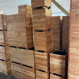 PAS-DOM SPÓŁKA Z OGRANICZONĄ ODPOWIEDZIALNOŚCIĄ - Magazyn pełen drewnianych donic skrzyniowych, ułożonych w stosy na betonowej posadzce, widoczne czarne wkłady w niektórych donicach.