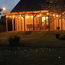 PAS-DOM SPÓŁKA Z OGRANICZONĄ ODPOWIEDZIALNOŚCIĄ - Oświetlona altana z drewnianą konstrukcją i meblami ogrodowymi, widoczna wieczorem z trawnika.