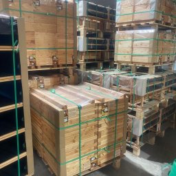 PAS-DOM SPÓŁKA Z OGRANICZONĄ ODPOWIEDZIALNOŚCIĄ - Stosy drewnianych elementów, prawdopodobnie desek lub belek, zapakowanych w folie i spiętych zielonymi taśmami, ułożone na paletach w magazynie.