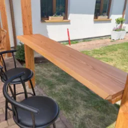 Zewnętrzny bar z drewnianym blatem, wsparty na drewnianej konstrukcji, z dwoma czarnymi krzesłami barowymi, na tle białego domu z oknami i zielonego trawnika.