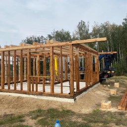 PAS-DOM SPÓŁKA Z OGRANICZONĄ ODPOWIEDZIALNOŚCIĄ - Konstrukcja drewniana domu w trakcie budowy, widoczny szkielet z belek na betonowej podstawie, w tle las i dźwig, na pierwszym planie butelka wody i zwinięty kabel.