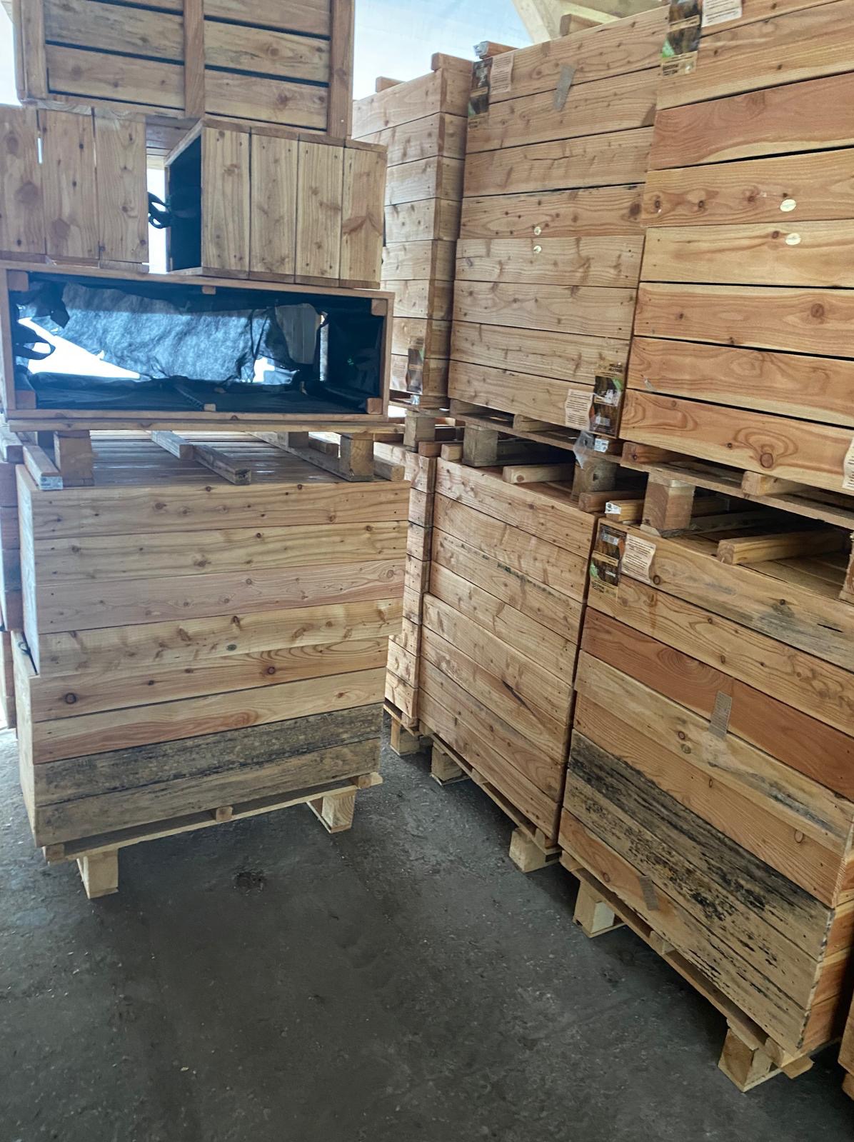 Stosy drewnianych skrzyń i palet w magazynie, niektóre skrzynie wyłożone czarną folią, widoczne ślady użytkowania i różnice w odcieniach drewna.