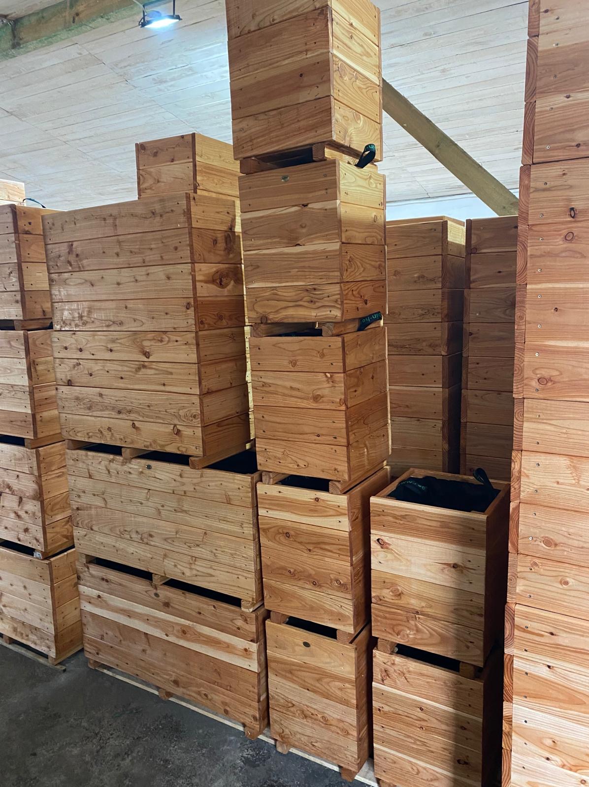 Magazyn pełen drewnianych donic skrzyniowych, ułożonych w stosy na betonowej posadzce, widoczne czarne wkłady w niektórych donicach.