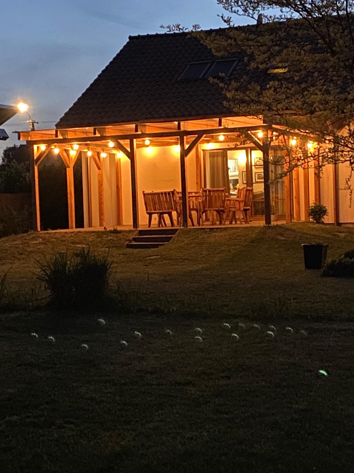 Oświetlona altana z drewnianą konstrukcją i meblami ogrodowymi, widoczna wieczorem z trawnika.