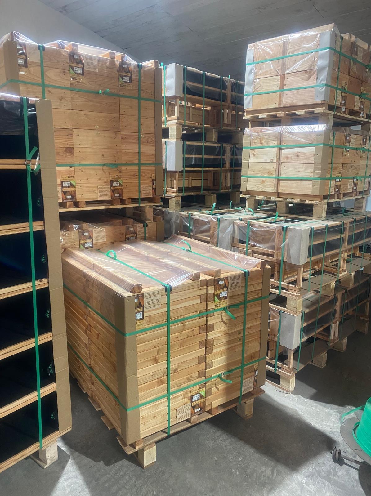 Stosy drewnianych elementów, prawdopodobnie desek lub belek, zapakowanych w folie i spiętych zielonymi taśmami, ułożone na paletach w magazynie.