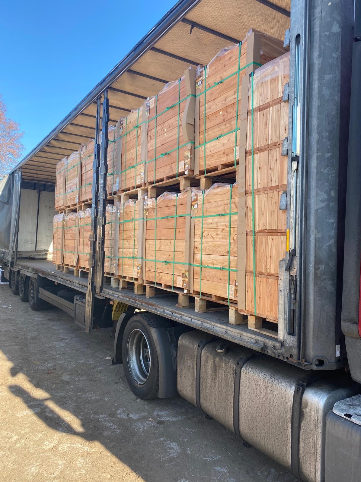 Ciężarówka z otwartą naczepą załadowana paletami z drewnianymi skrzyniami, zabezpieczonymi zielonymi taśmami, widok z boku na tle błękitnego nieba.