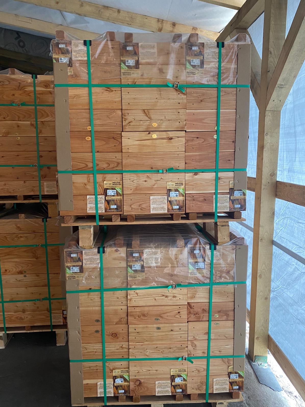 Palety z drewnianymi skrzynkami, owinięte folią i spięte zielonymi pasami transportowymi, składowane w zadaszonej przestrzeni z drewnianą konstrukcją nośną.