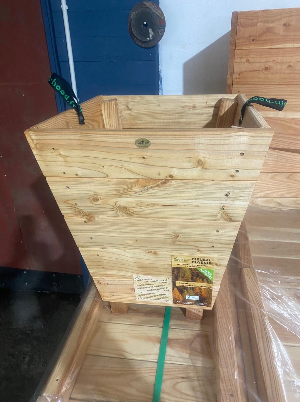 Drewniana, trapezoidalna donica z jasnego drewna, z widocznymi sękami i etykietą informacyjną, ustawiona w drewnianej skrzyni transportowej, z elementami mocującymi i napisem 'hood.fr'.