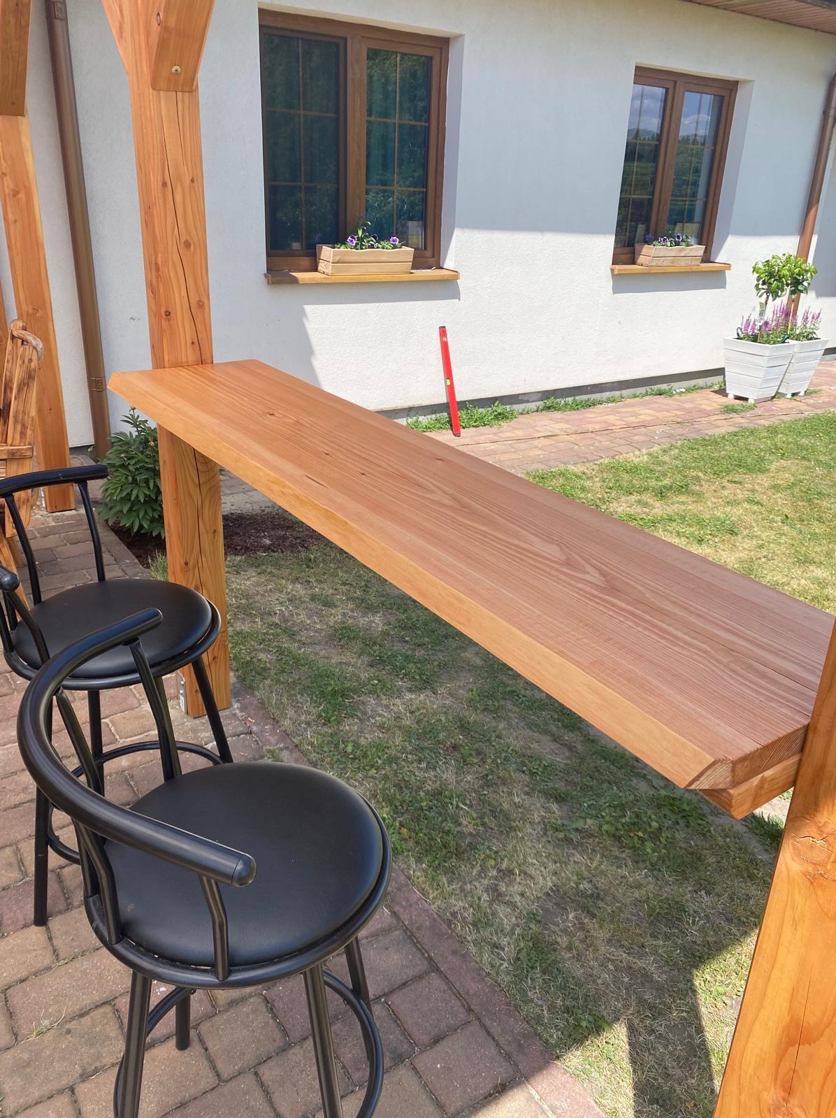 Zewnętrzny bar z drewnianym blatem, wsparty na drewnianej konstrukcji, z dwoma czarnymi krzesłami barowymi, na tle białego domu z oknami i zielonego trawnika.