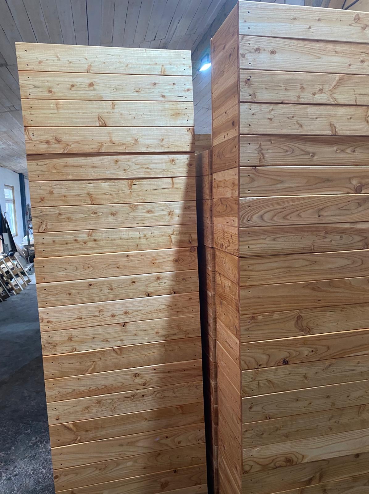 Panele z jasnego drewna ułożone w stos, widoczne sęki i usłojenie, zarys palet w tle.