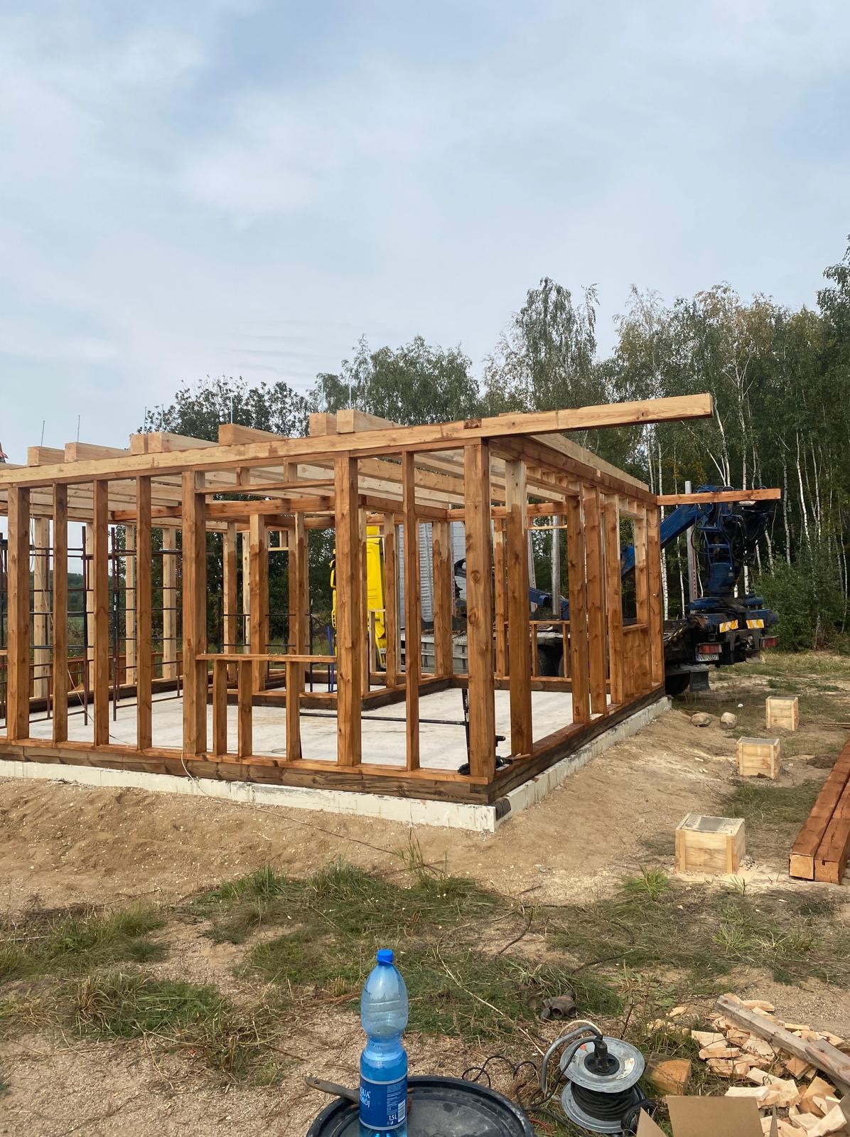 Konstrukcja drewniana domu w trakcie budowy, widoczny szkielet z belek na betonowej podstawie, w tle las i dźwig, na pierwszym planie butelka wody i zwinięty kabel.