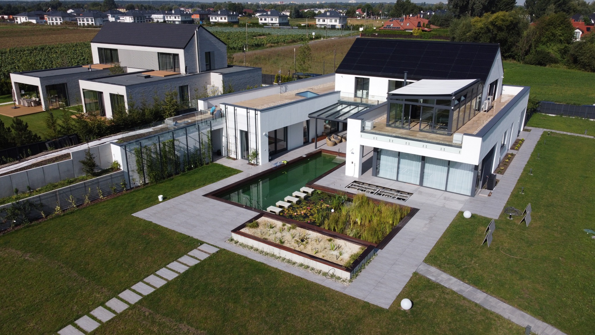 Nowoczesny dom z basenem i panelami słonecznymi w Warszawie, widok z lotu ptaka. Minimalistyczna architektura, zadbany ogród i elementy wodne.