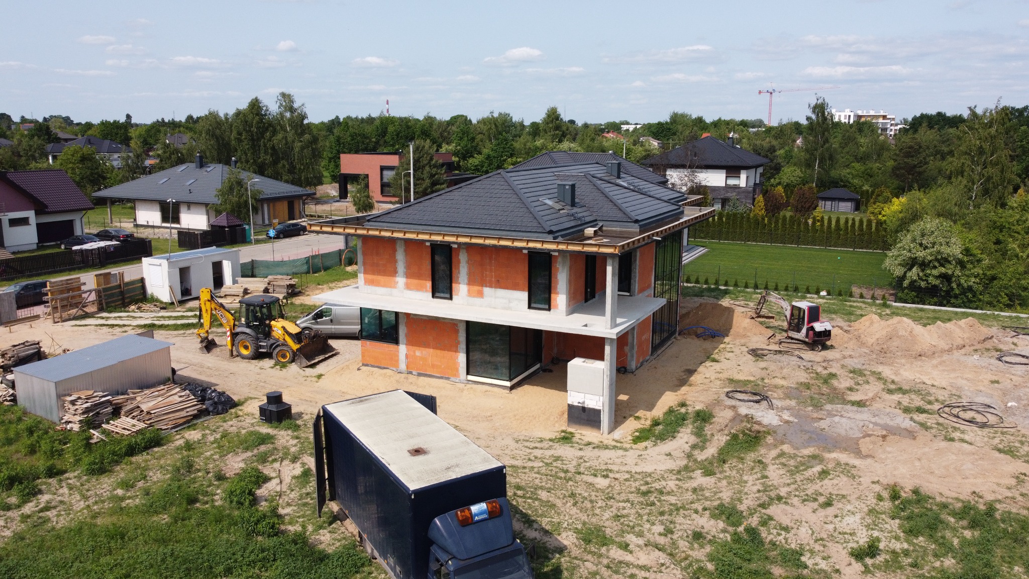 Dwukondygnacyjny dom w trakcie budowy z pomarańczowej cegły, z czarnym dachem i dużymi oknami. Na posesji sprzęt budowlany i ciężarówka dostawcza. Widok z góry.