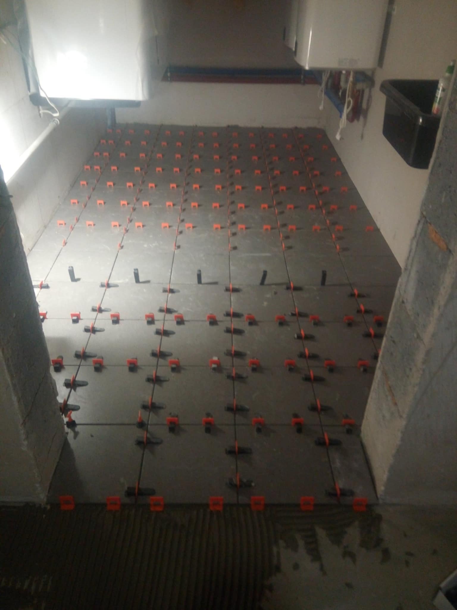 Szare płytki podłogowe w trakcie układania, z pomarańczowymi klipsami poziomującymi na fugach, widoczne rury i zbiornik na ścianie w tle.