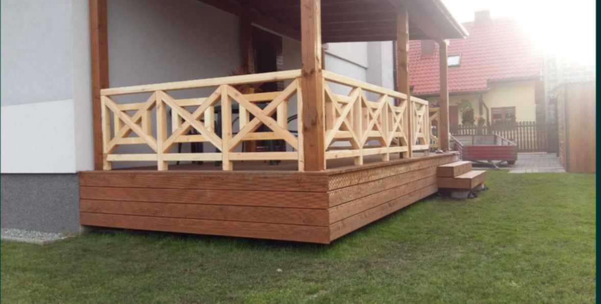 Zadaszony taras z drewnianą balustradą o geometrycznym wzorze, brązowa podłoga z desek, widok na zielony trawnik i dom z czerwoną dachówką w tle.