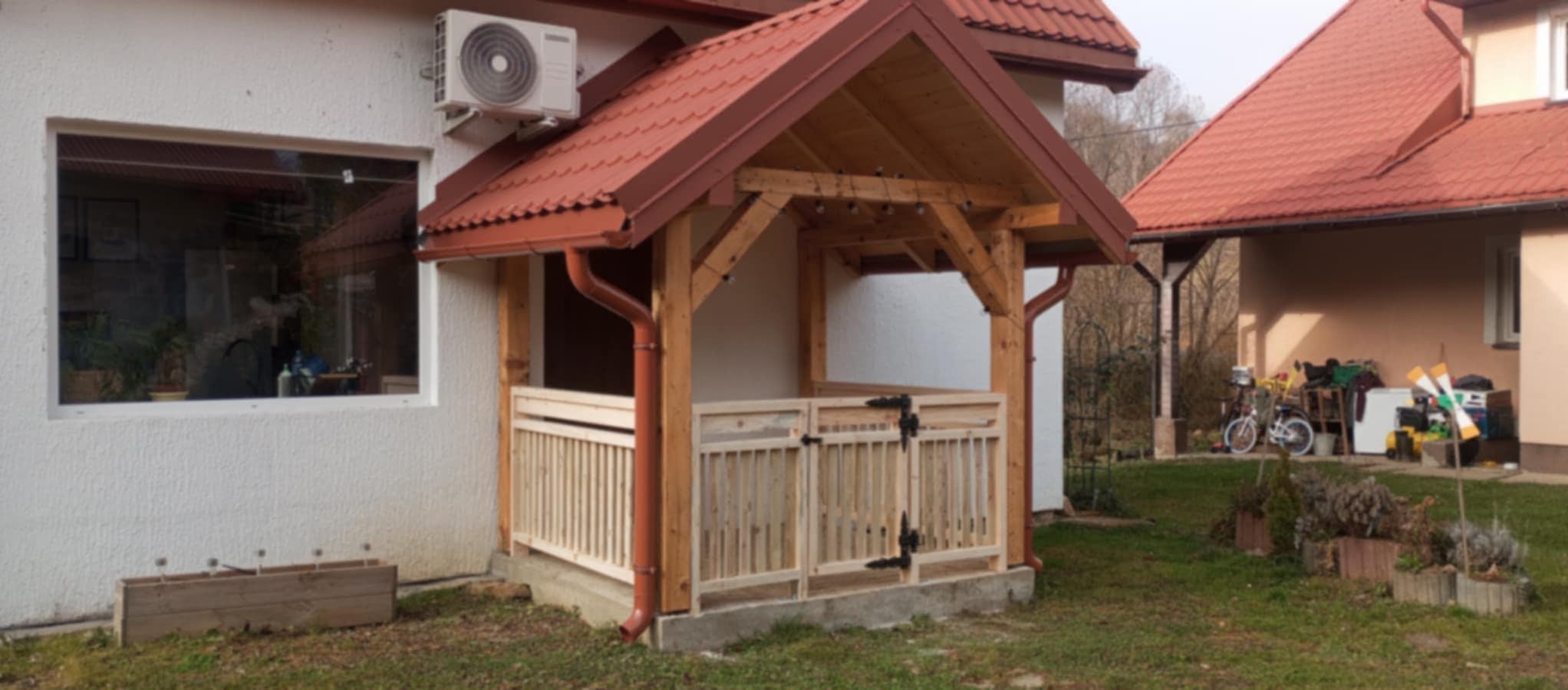 Drewniana weranda z brązowym dachem i rynnami, przymocowana do białego domu z klimatyzatorem, z widocznymi elementami ogrodzenia i skrzynią na kwiaty.