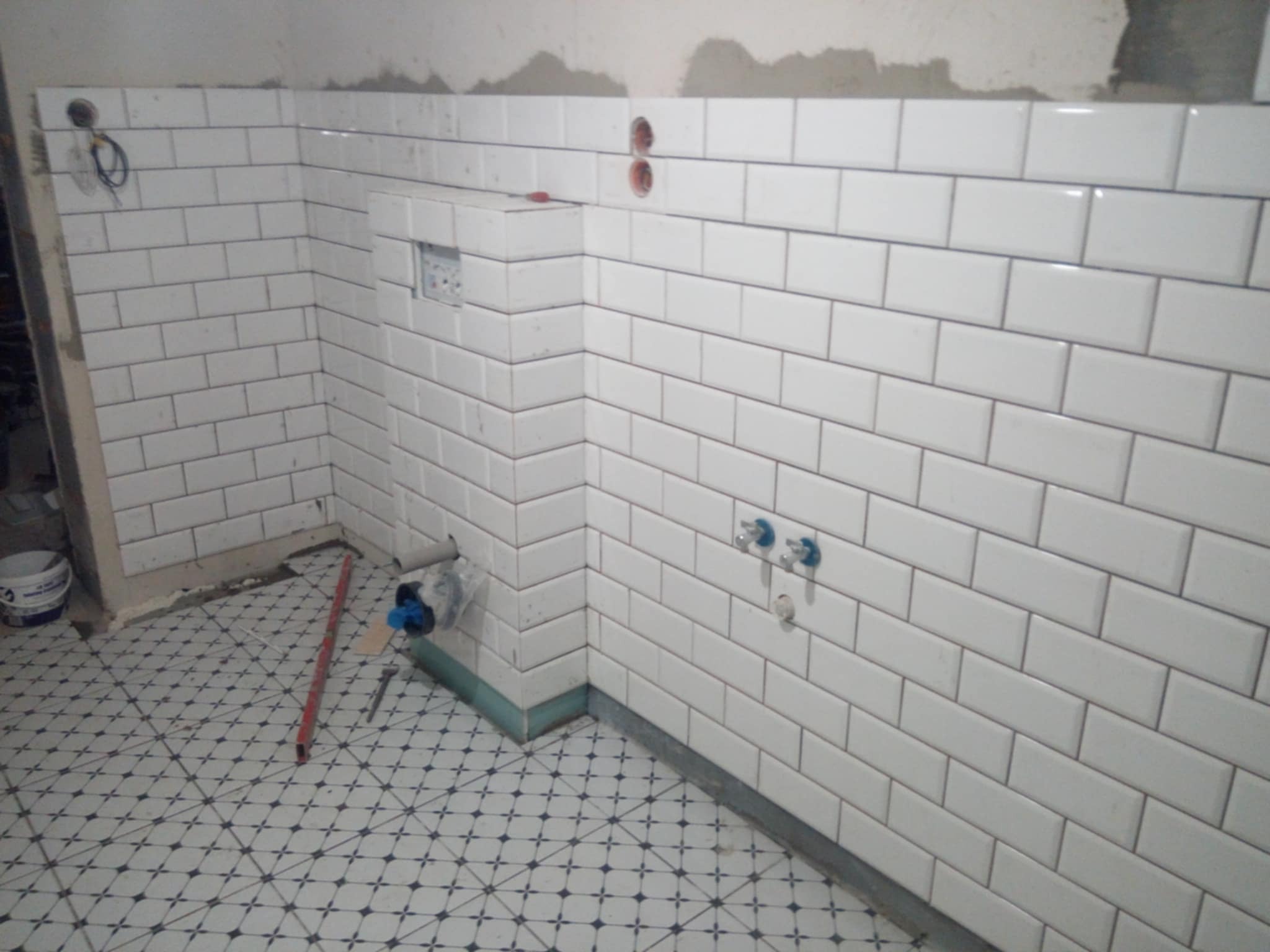 Ściana w trakcie wykańczania z białymi, prostokątnymi płytkami ułożonymi w cegiełkę, widoczne instalacje hydrauliczne i elektryczne, podłoga z wzorem geometrycznym w kolorze biało-czarnym...