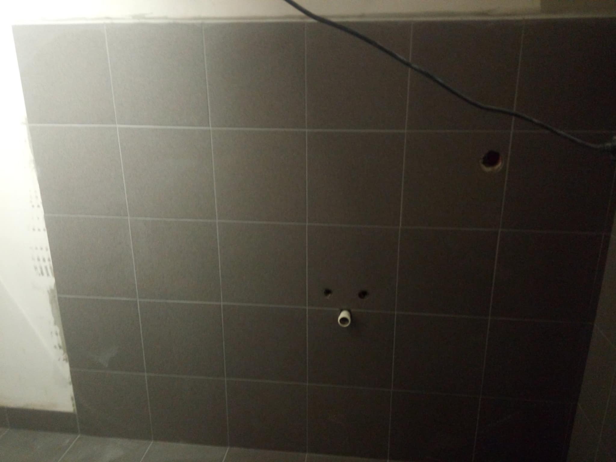 Ściana w łazience wyłożona ciemnymi, kwadratowymi płytkami. Widoczne otwory na rury i instalację. Po lewej stronie fragment ściany przygotowanej pod płytki.