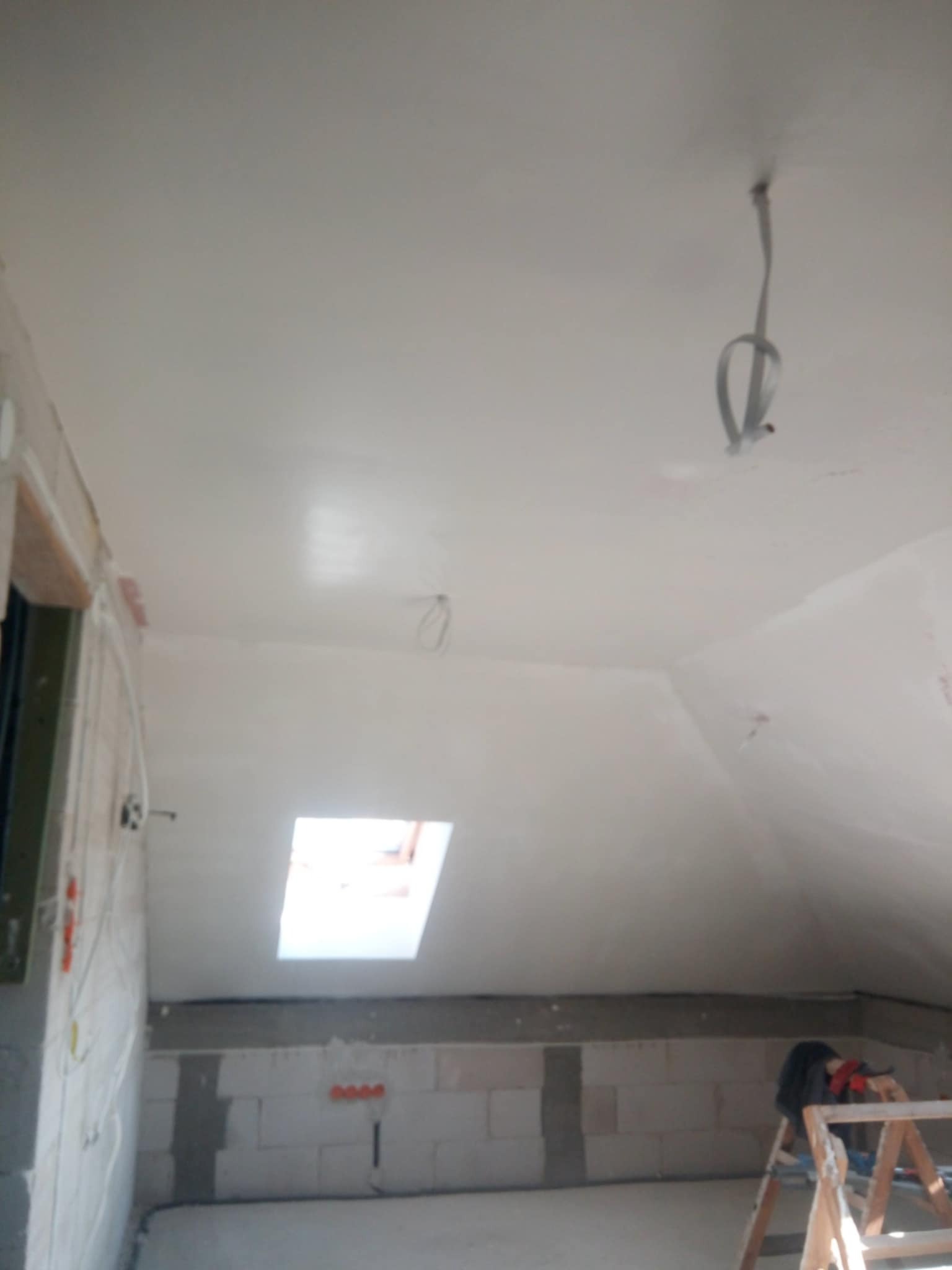 Poddasze w trakcie prac wykończeniowych; ściany i skośny sufit pokryte świeżo nałożoną, jasną gładzią; widoczne niedoskonałości i przewody wystające z sufitu; na dole ściana z bloczków betonowych...