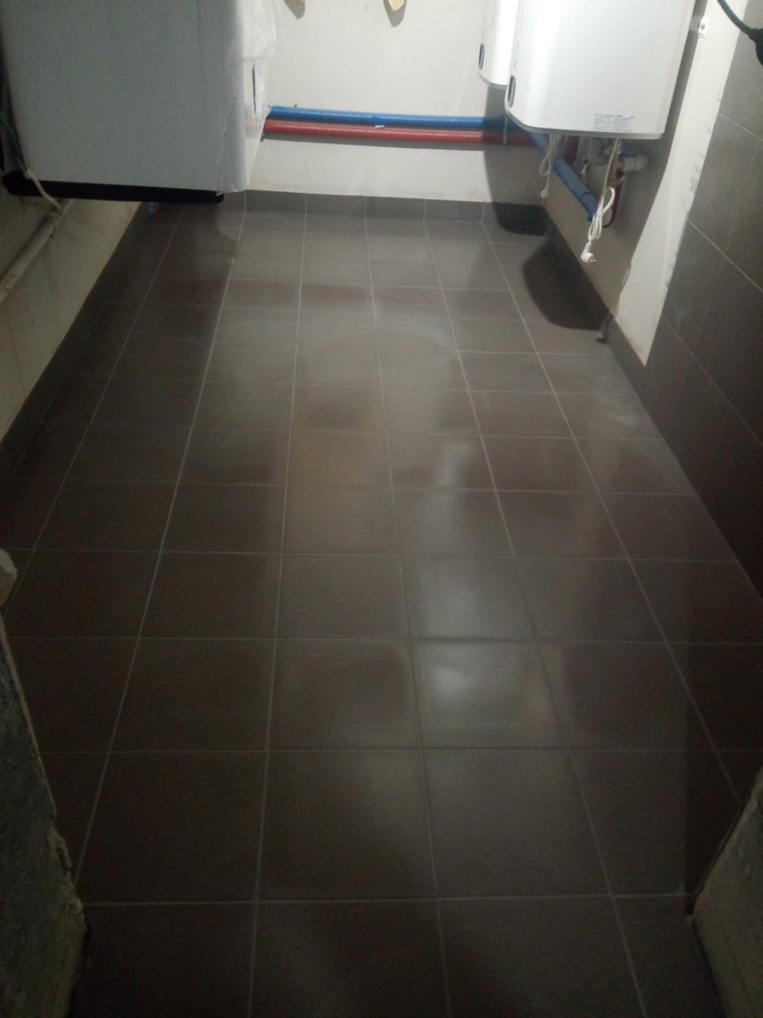 Nowo ułożona, ciemnobrązowa podłoga z płytek w pomieszczeniu technicznym, widoczne rury instalacyjne na ścianie i białe zasobniki ciepłej wody.