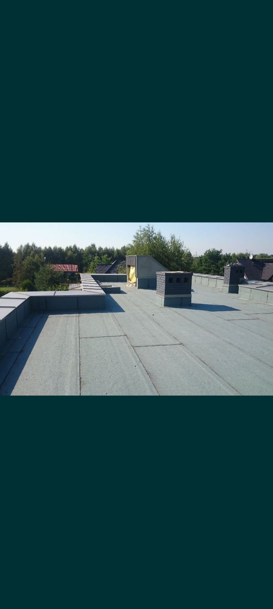 Płaski dach pokryty papą termozgrzewalną, widoczne kominy wentylacyjne i obróbki blacharskie, w tle drzewa.