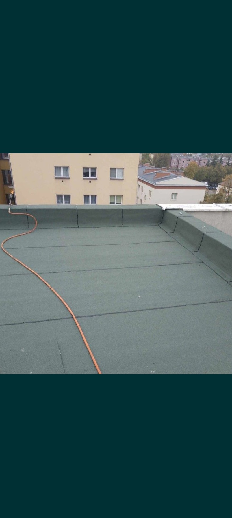 Zielony dach płaski z widocznymi łączeniami papy bitumicznej, w tle budynki mieszkalne i pomarańczowy wąż.