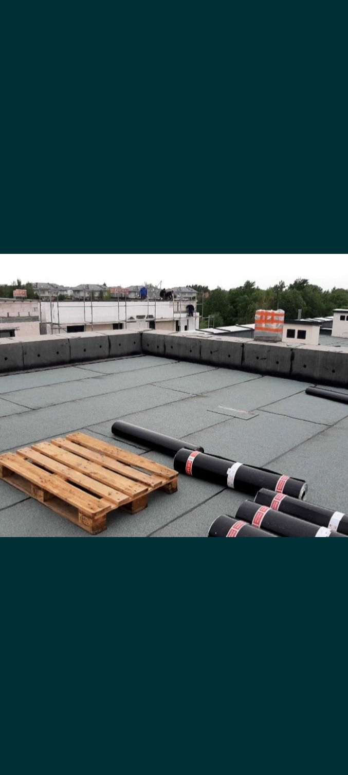 Wykończony płaski dach z papy termozgrzewalnej, widoczne rolki papy na palecie, w tle ekipa montująca obróbki blacharskie.